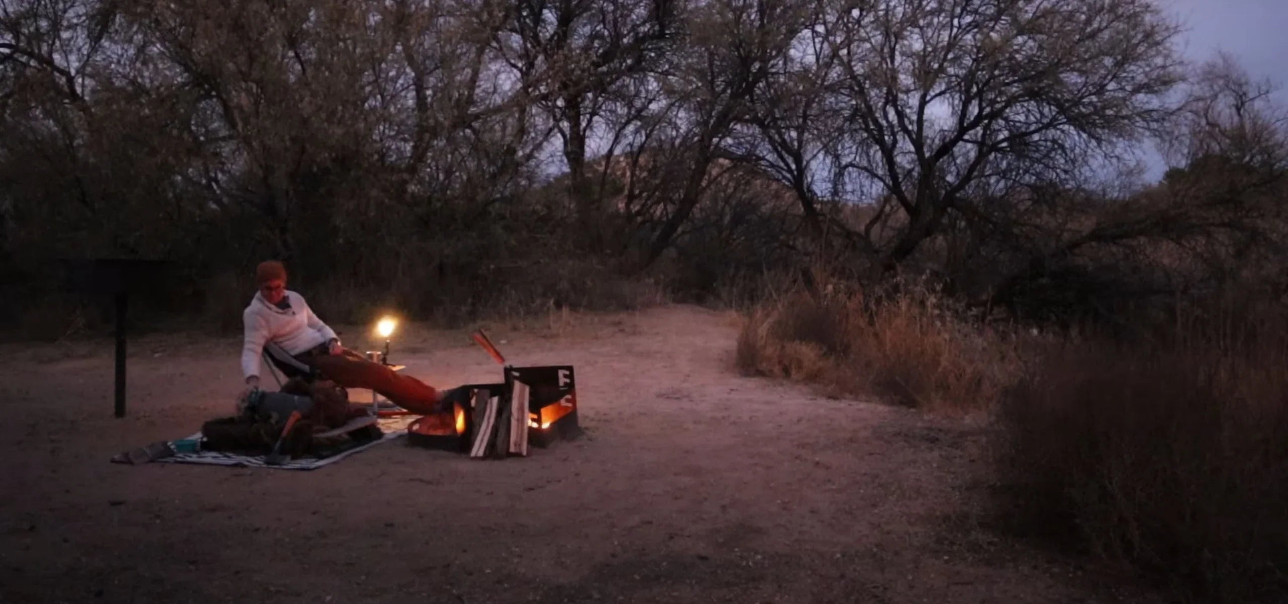 Camper relaxing by campfire with kettle pot and gear at dusk in a wooded wilderness campsite