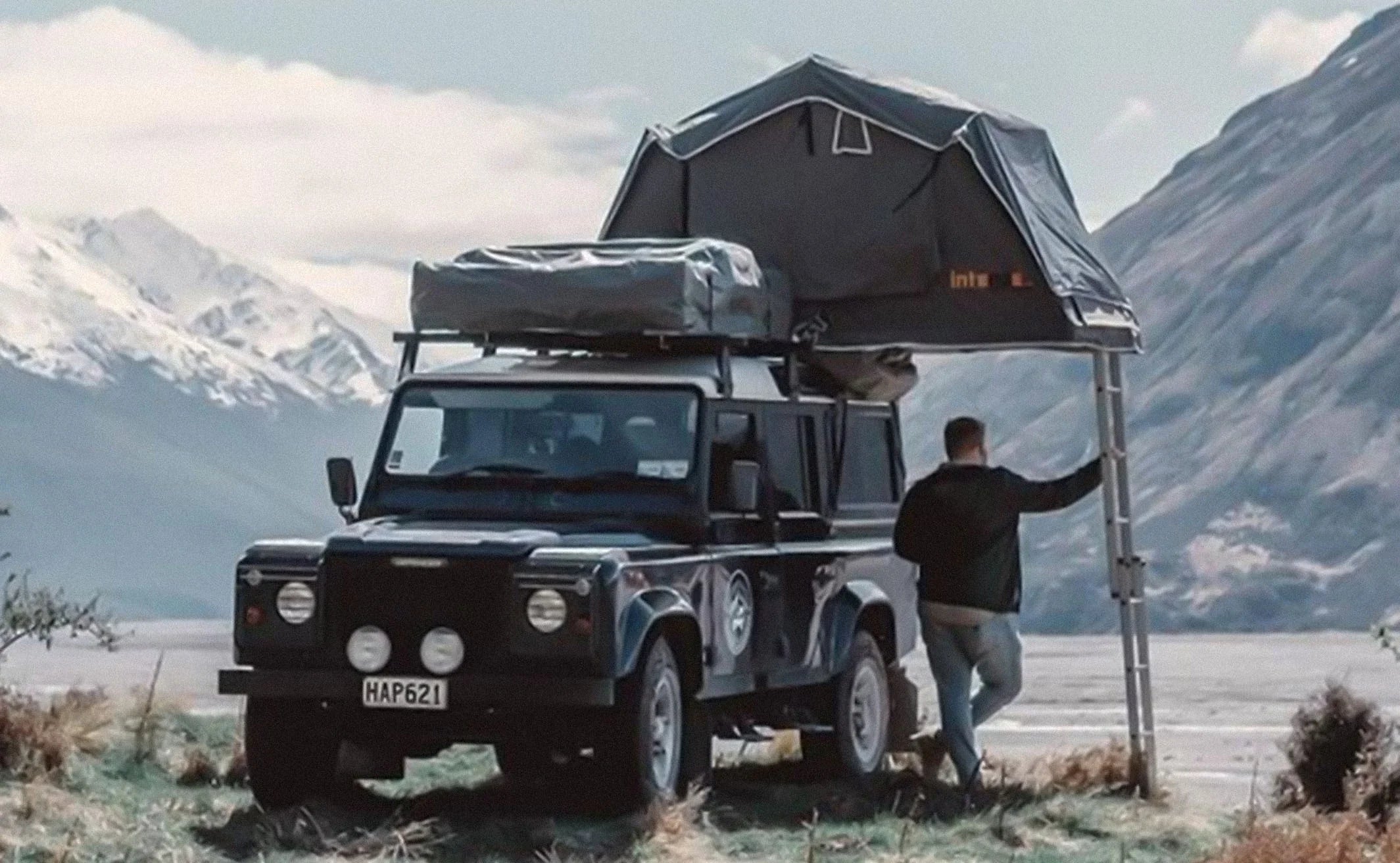 Man camping with off-road SUV and rooftop tent by mountains and river, outdoor adventure scene